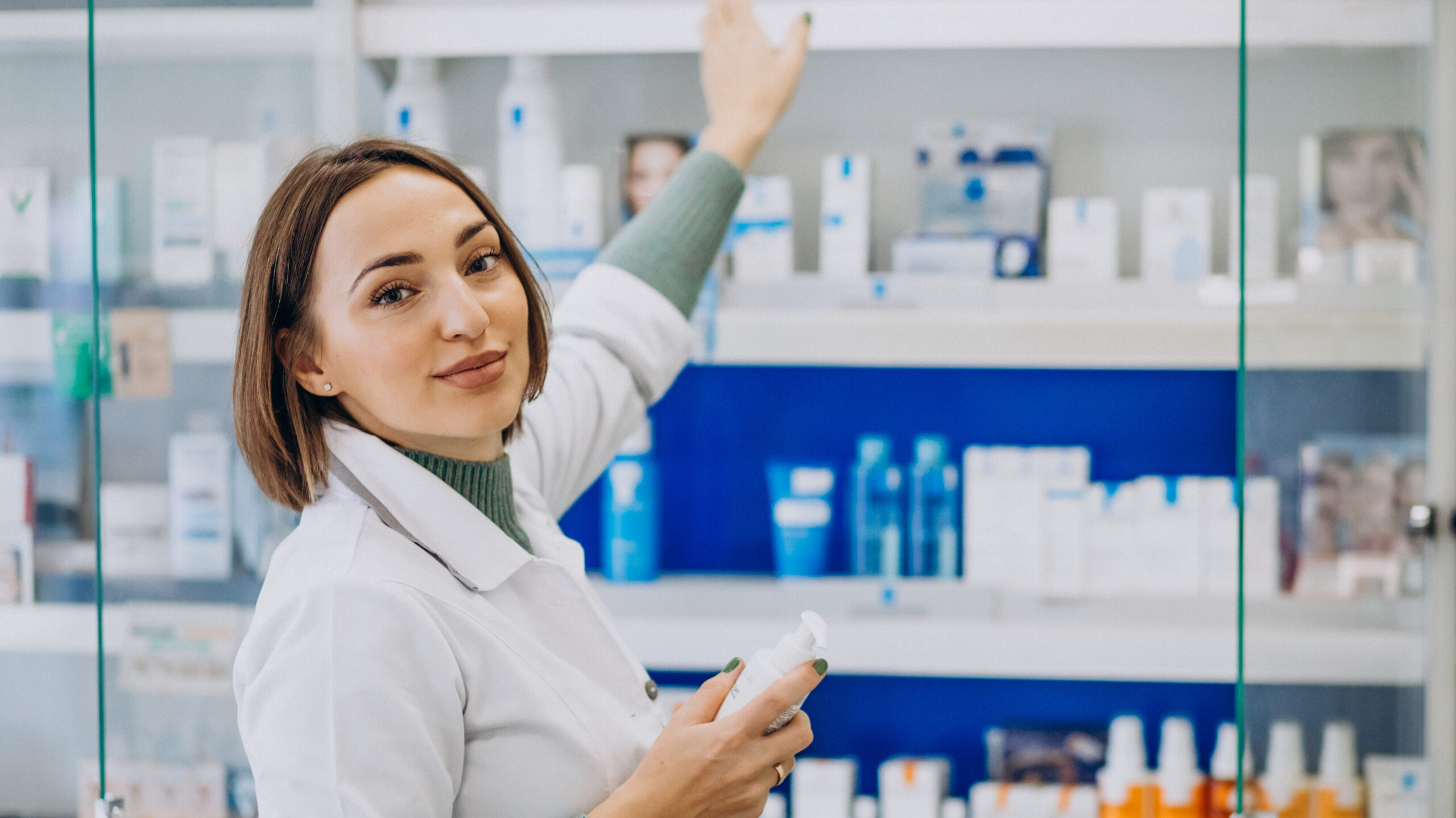 Mujer joven con cabello castaño viste una bata blanca y extiende su mano hacia un estante de medicinas en farmacia
